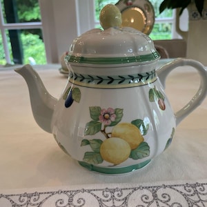 May include: A white ceramic teapot with a matching lid featuring a fluted design. The body is decorated with painted yellow lemons, green leaves, and small pink flowers. A green and yellow border pattern encircles the top and base of the teapot.