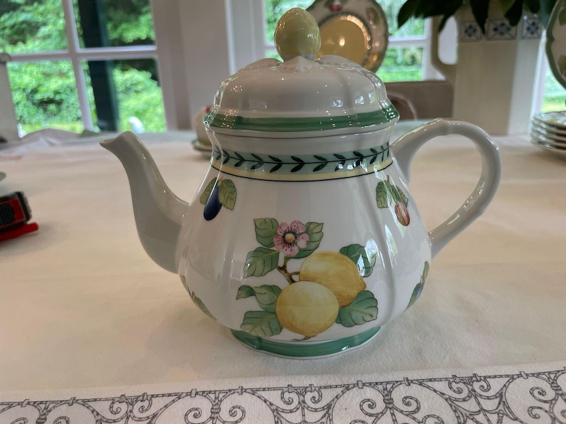 May include: A white ceramic teapot with a matching lid featuring a fluted design. The body is decorated with painted yellow lemons, green leaves, and small pink flowers. A green and yellow border pattern encircles the top and base of the teapot.