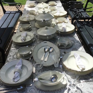 May include: An outdoor dining table set with cream-coloured porcelain tableware. The arrangement includes plates, bowls, cups, and serving dishes, complemented by silverware, crystal glasses, and folded napkins. The table is set on a white lace tablecloth.