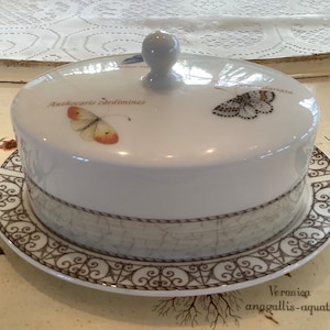 Op de afbeelding: Witte keramische boterpot met een deksel met vlinderillustraties en botanische tekst. De schaal staat op een bijpassend bord met een decoratieve rand. Het deksel heeft een kleine, ronde handgreep.