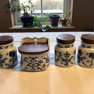 May include: Four white ceramic spice jars with blue floral designs and wooden lids. The jars are arranged in a row on a white table. One jar is a wall-mounted spice rack with a hinged lid.