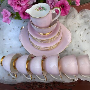 May include: A set of pink teacups and saucers with gold trim. The cups and saucers are stacked on top of each other. There is also a pink creamer with gold trim.