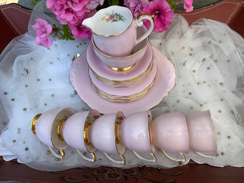 May include: A set of pink teacups and saucers with gold trim. The cups and saucers are stacked on top of each other. There is also a pink creamer with gold trim.