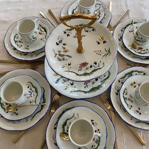 May include: A two-tiered serving stand with a gold handle, decorated with floral patterns in shades of blue, green, and yellow. The stand is surrounded by six white plates with blue trim, each holding a small white teacup with a floral design.