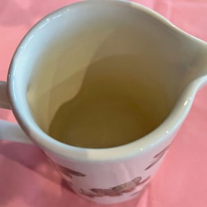 Puede incluir: Una jarra de leche de cer&aacute;mica blanca con interior liso y pico vertedor. El exterior presenta un patr&oacute;n floral pintado en tonos marrones y rojos. Un asa curva est&aacute; unida al lateral, mostrando un acento de pintura roja en la parte superior.
