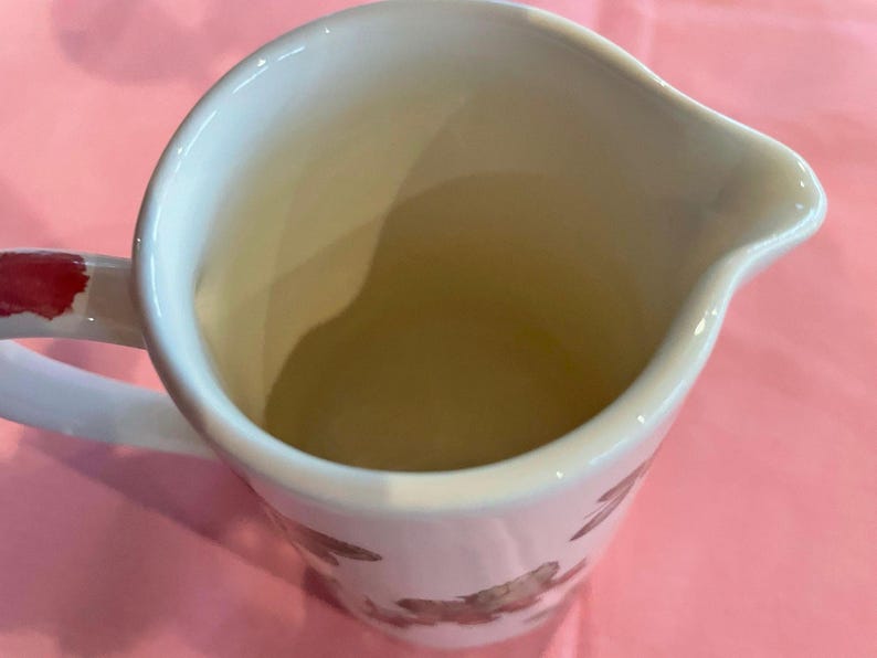 Puede incluir: Una jarra de leche de cer&aacute;mica blanca con interior liso y pico vertedor. El exterior presenta un patr&oacute;n floral pintado en tonos marrones y rojos. Un asa curva est&aacute; unida al lateral, mostrando un acento de pintura roja en la parte superior.