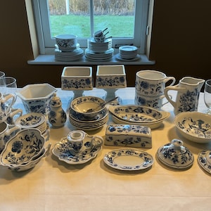 Può includere: Una collezione di stoviglie in porcellana blu e bianca, tra cui tazze, piattini, ciotole, piatti e vassoi. I pezzi presentano un motivo floreale e sono disposti su una tovaglia bianca. Sono presenti varie dimensioni e forme.