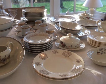 May include: A white table setting with a variety of plates, bowls, and cups. The dishes are decorated with gold trim and floral patterns. There are also a few serving dishes and a gravy boat.