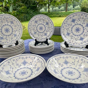 May include: A collection of white ceramic dinnerware featuring a blue floral pattern with intricate scrolling vines and central medallions. The set includes several stacked plates and two shallow bowls, all decorated with a consistent blue design.