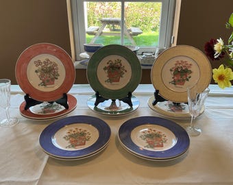 Op de afbeelding: Een set van vijf keramische borden met gekleurde randen in koraal, groen, geel en blauw. Elk bord toont een centrale botanische illustratie van bloemen in een pot. Twee heldere glazen bekers staan op het witte tafelkleed.