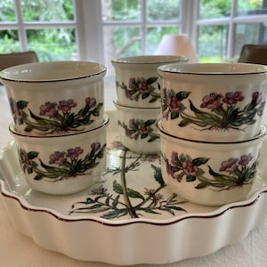 May include: A set of six white ceramic ramekins with floral designs. The ramekins are stacked in pairs and arranged on a white ceramic serving dish with a scalloped edge.