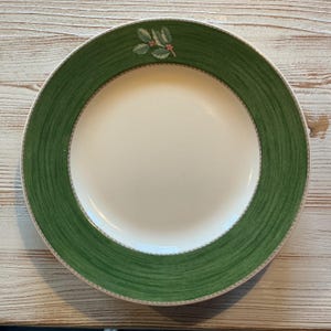 Puede incluir: Un plato redondo de cerámica con un centro blanco sólido y un borde verde ancho y texturizado. El borde verde presenta un patrón sutil similar a pinceladas y una pequeña ilustración detallada de hojas de acebo verdes con bayas rojas.