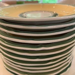 May include: A stack of ceramic saucers featuring a pale yellow centre, a white rim, and a dark green band along the edge. Each saucer has a scalloped, wavy border design. The top saucer displays a dark green laurel leaf pattern in the centre.