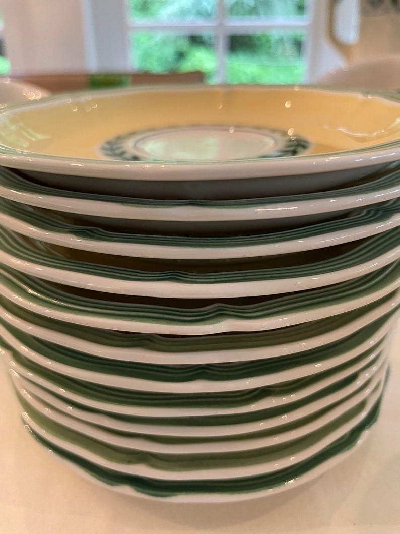 May include: A stack of ceramic saucers featuring a pale yellow centre, a white rim, and a dark green band along the edge. Each saucer has a scalloped, wavy border design. The top saucer displays a dark green laurel leaf pattern in the centre.