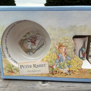 Peut inclure: Un coffret Peter Rabbit par Wedgwood contenant un bol et une tasse en céramique. Le bol présente une illustration de lapin avec un panier, et la tasse montre un lapin en veste bleue. La boîte affiche du texte et une scène de jardin.