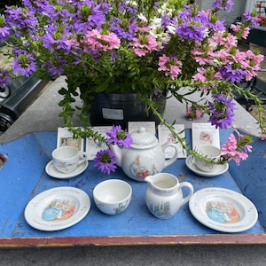 Può includere: Un vassoio di legno blu con un manico su ogni lato, che contiene un set da tè Peter Rabbit. Il set comprende una teiera, un creamer, uno zuccheriera e due tazze da tè e piattini. Il vassoio è decorato con un'arrangiamento floreale di fiori viola e rosa.