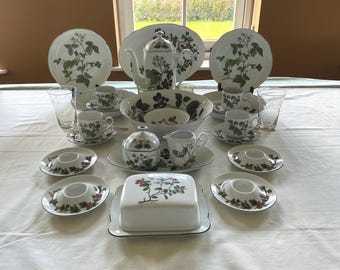 May include: A complete white porcelain tea set with a botanical design featuring green leaves and red berries. The set includes plates, cups, saucers, a teapot, a sugar bowl, a creamer, and serving dishes. The set is arranged on a white tablecloth.