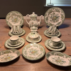 Puede incluir: Un juego de té completo con un estampado floral en verde, amarillo y blanco. El juego incluye tazas de té, platillos, platos, una tetera, una azucarera y fuentes. Los artículos están colocados sobre una mesa de madera.