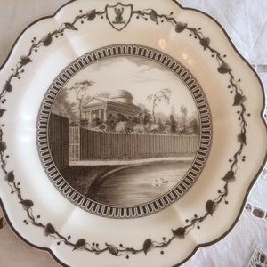 Può includere: Un piatto in ceramica bianca con un design marrone e bianco. Il piatto presenta un'illustrazione in bianco e nero di un edificio con una recinzione e alberi. Il piatto ha un bordo ondulato e un design floreale marrone attorno al bordo.