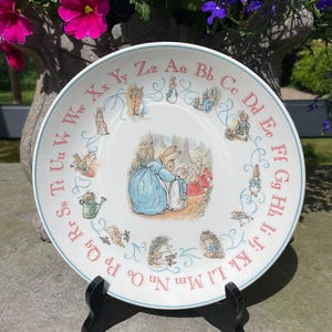 Puede incluir: Un plato de cerámica blanco con un borde azul presenta una ilustración de Peter Rabbit en el centro. El plato está decorado con el alfabeto alrededor del borde, con cada letra en un color diferente.