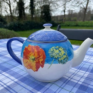 May include: A ceramic teapot with a blue handle and lid, featuring a floral design. The teapot has a white body with a large orange and yellow flower and a yellow and blue flower painted on the side. The lid is blue with a white knob.