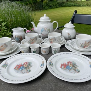 May include: A white ceramic tea set featuring illustrations of Peter Rabbit. The set includes a teapot, sugar bowl, creamer, four cups with saucers, four small egg cups, and two plates, all decorated with colorful scenes and text from the story.