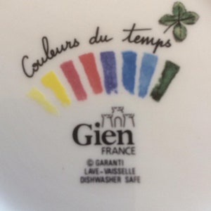 Puede incluir: Una superficie de cer&aacute;mica blanca con el texto Couleurs du temps en letra cursiva negra, un tr&eacute;bol de cuatro hojas y un arco&iacute;ris de pinceladas amarillas, rojas, moradas, azules y verdes. Debajo, el logotipo de Gien France y Dishwasher Safe.