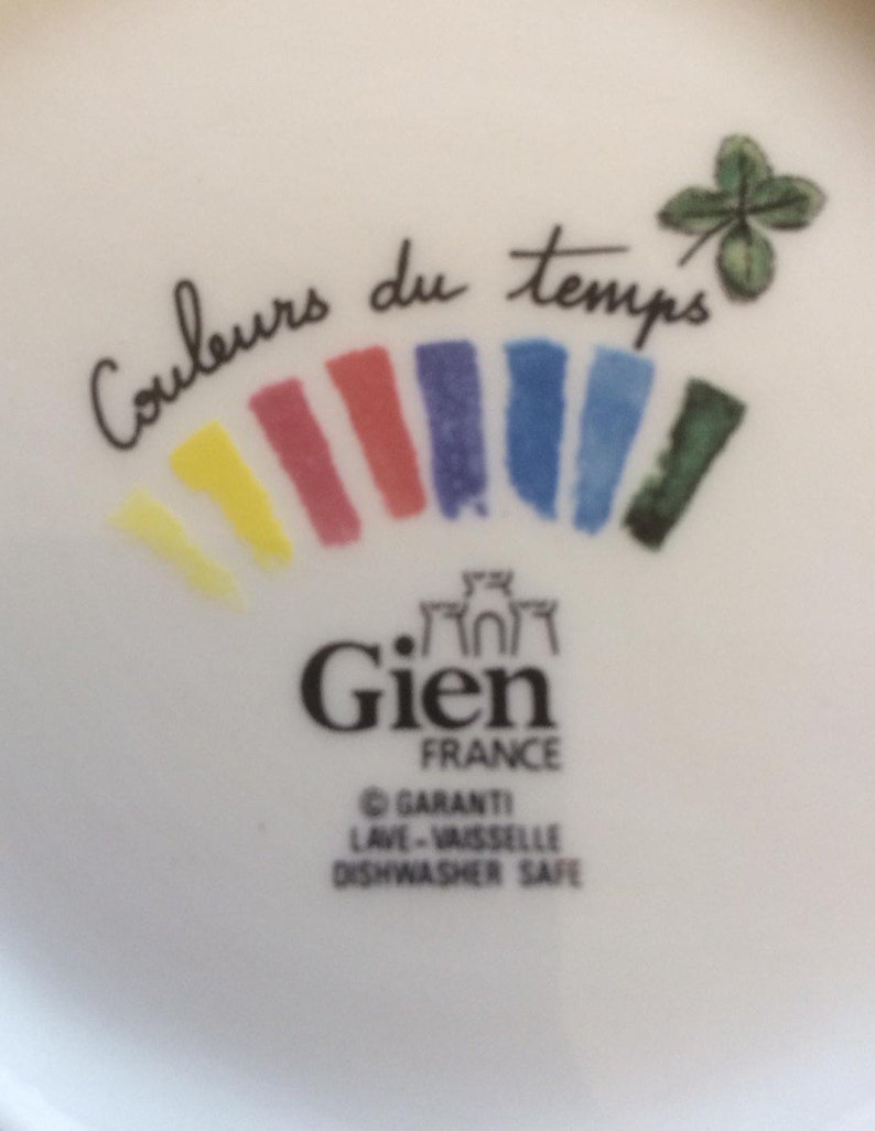 Puede incluir: Una superficie de cer&aacute;mica blanca con el texto Couleurs du temps en letra cursiva negra, un tr&eacute;bol de cuatro hojas y un arco&iacute;ris de pinceladas amarillas, rojas, moradas, azules y verdes. Debajo, el logotipo de Gien France y Dishwasher Safe.
