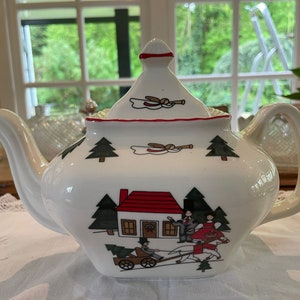 May include: White ceramic teapot with a red and green Christmas scene. The teapot has a lid with a red trim and a white handle. The scene features a small red house with a white door and windows, two green trees, and a horse-drawn carriage with two figures.