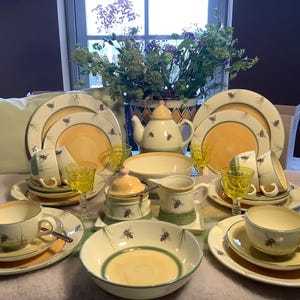 Może przedstawiać: Ceramiczny zestaw obiadowy w kolorach kremowym, żółtym i zielonym z motywami pszczół i igieł sosnowych. Kolekcja zawiera talerze, miski, filiżanki, spodki, dzbanek do herbaty, dzbanek na mleko, cukiernicę oraz żółte kieliszki.