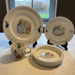 Peut inclure: Un ensemble de vaisselle en céramique orné d'illustrations de Pierre Lapin et de texte. Il comprend une grande assiette, une petite assiette, une tasse et un bol peu profond, décorés de scènes colorées de lapins et de texte narratif imprimé.