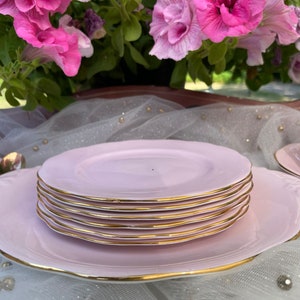 May include: A set of pink teacups and saucers with gold trim. The teacups and saucers are stacked on top of each other. The teacups and saucers are on a white tablecloth.