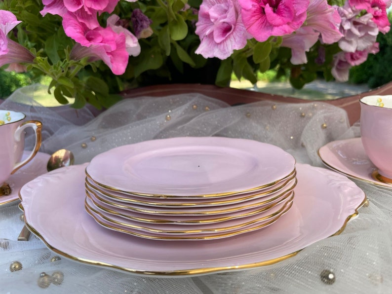 May include: A set of pink teacups and saucers with gold trim. The teacups and saucers are stacked on top of each other. The teacups and saucers are on a white tablecloth.