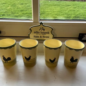以下が含まれることがあります： 緑色のスカラップリムと黒い雄鶏のデザインが施された、4つの黄色のセラミックグラス。グラスには「zeller keramik Hahn & Henne」と表示されています。グラスは、緑の草の背景がある白い窓枠にあります。