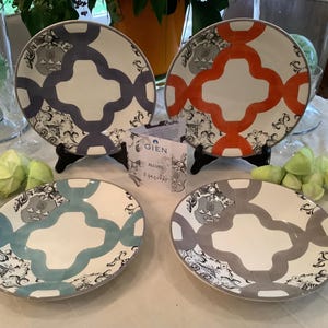 Puede incluir: Cuatro platos de cena de cerámica con fondo blanco y un patrón geométrico en tonos de azul, gris, naranja y verde azulado. Los platos están dispuestos sobre una mesa blanca con hojas verdes y una tarjeta que dice "Gien Allieur."