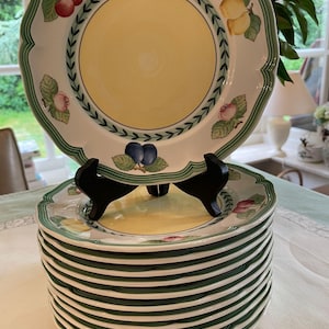 May include: A stack of white ceramic plates with a scalloped edge and a central yellow circle. The rim features a green border with a leaf pattern and illustrations of cherries, lemons, and plums. The top plate rests on a black plastic stand.