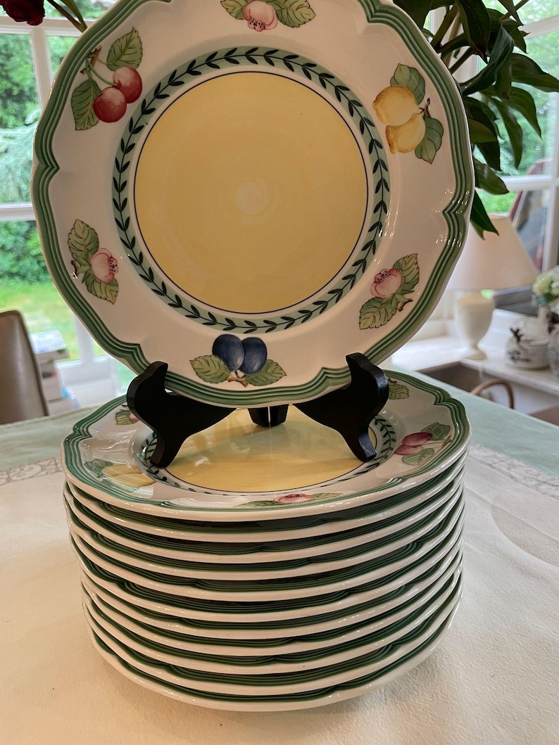 May include: A stack of white ceramic plates with a scalloped edge and a central yellow circle. The rim features a green border with a leaf pattern and illustrations of cherries, lemons, and plums. The top plate rests on a black plastic stand.