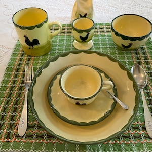 May include: A table setting with a green and yellow checkered tablecloth. There are two mugs, a bowl, a plate, a cup and saucer, a spoon, a fork, and an egg cup, all with a rooster design.
