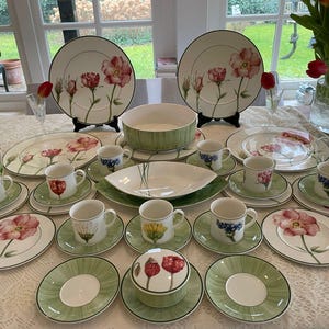 Może przedstawiać: Kompletny zestaw naczyń o tematyce kwiatowej, składający się z talerzy, misek, filiżanek i spodków. Zestaw zawiera dużą miskę do serwowania i zakryty pojemnik. Każdy element zdobi różowy kwiatowy wzór i zielone akcenty.