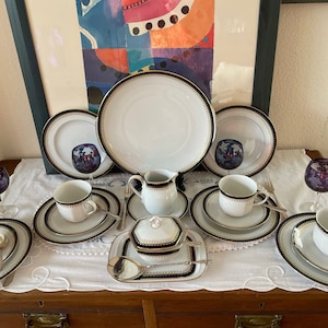 Könnte beinhalten: Ein Set aus weißen Porzellan-Teetassen und Untertassen mit schwarzem und goldenem Rand. Das Set beinhaltet eine Teekanne, eine Zuckerdose und eine Milchkännchen. Die Tassen und Untertassen haben ein zartes Blumenmuster.