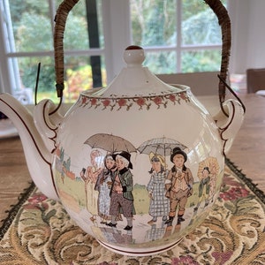 Op de afbeelding: Een witte theepot van keramiek met een bruine rieten handvat. De theepot heeft een fantasievolle illustratie van kinderen die met paraplu's in de regen lopen. De theepot heeft een bloemenmotief rond de bovenkant.
