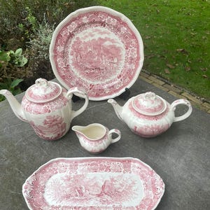 Puede incluir: Un conjunto de vajilla de cerámica rosa y blanca con un diseño de paisaje. El conjunto incluye un plato grande, dos teteras, una jarra de crema y una fuente para servir. El plato tiene aproximadamente 25 cm de diámetro.