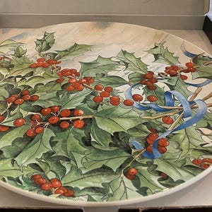 May include: A round ceramic plate featuring a detailed illustration of green holly leaves and bright red berries. A thin blue ribbon is intertwined among the branches. The plate is presented inside an open black and white cardboard box.