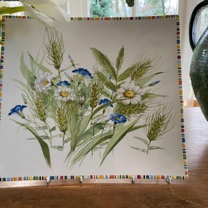 Peut inclure: Une assiette carrée décorative avec un motif floral de marguerites blanches, de fleurs bleues et d'épis de blé. L'assiette a une bordure rayée colorée. L'assiette est posée sur une surface en bois.