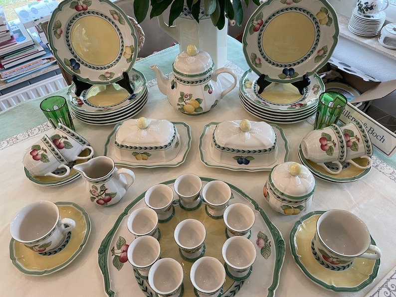 May include: A Villeroy & Boch French Garden dinnerware set featuring white ceramic pieces with yellow centres and green rims decorated with fruit motifs. The collection includes plates, a teapot, lidded butter dishes, cups, saucers, and egg cups.