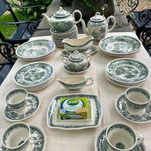 Puede incluir: Un juego de vajilla de cerámica con un patrón pastoral de transferencia en verde y blanco. La colección incluye teteras, tazas de té con platillos, platos, salseras, un azucarero y un plato cuadrado con una caja de mantequilla Campina.