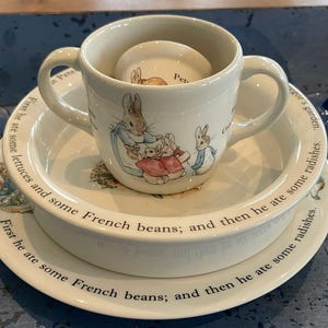 May include: A cream-coloured ceramic two-handled mug, bowl, and plate set featuring illustrations of Peter Rabbit. The pieces are decorated with text reading, First he ate some lettuces and some French beans; and then he ate some radishes.
