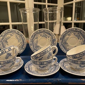 May include: A set of white teacups and saucers with blue floral designs, displayed on a blue tray. Two tall, clear glass vases are in the background. The teacups are stacked, and the saucers are arranged around them.