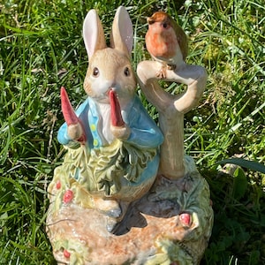 Puede incluir: Una figurita de cerámica de Peter Rabbit sosteniendo dos zanahorias, de pie sobre una base con hojas verdes y bayas rojas. Un petirrojo está posado en un pequeño árbol detrás de Peter Rabbit.