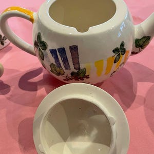 Puede incluir: Una tetera de cer&aacute;mica blanca con tapa a juego, que presenta un dise&ntilde;o pintado a mano de hojas verdes, bayas rojas y patrones de pinceladas verticales en amarillo y azul. El asa tiene un detalle amarillo y la tetera tiene una abertura superior.